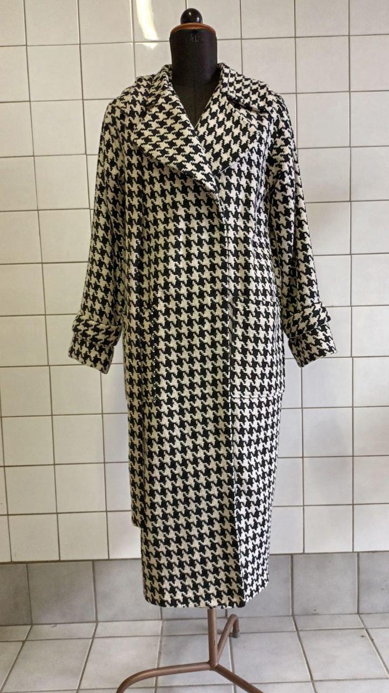 May include: A black and white houndstooth pattern coat displayed on a mannequin. The long coat has a notched collar, long sleeves, and pockets. The coat is knee-length and has a classic, timeless design.