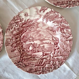 Può includere: Un piatto rotondo in ceramica con bordo smerlato caratterizzato da una stampa transfer color marrone scuro e bianco. Il disegno raffigura un paesaggio rurale con un cacciatore che punta un fucile, un cane, un mulino ad acqua, alberi e un uccello.