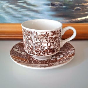 Könnte beinhalten: Eine weiße Teetasse mit passender Untertasse aus Keramik, verziert mit einem detaillierten braunen Transferdruckmuster. Das Design zeigt eine historische Straßenszene mit einer Pferdekutsche, Fachwerkhäusern und Figuren in historischer Kleidung.