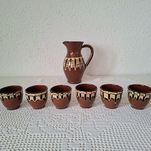 Könnte beinhalten: Ein brauner Keramikkrug und sechs passende Tassen. Der Krug hat einen Henkel und ein dekoratives Muster aus schwarzen und gelben Linien. Die Tassen haben ein ähnliches Design am Rand. Das Set wird auf einer weißen, strukturierten Oberfläche präsentiert.