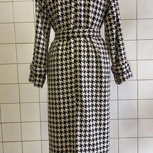 May include: A long, houndstooth-patterned coat in black and cream. The coat features a belted waist, long sleeves with buttoned cuffs, and a classic collar. The coat is displayed on a mannequin.