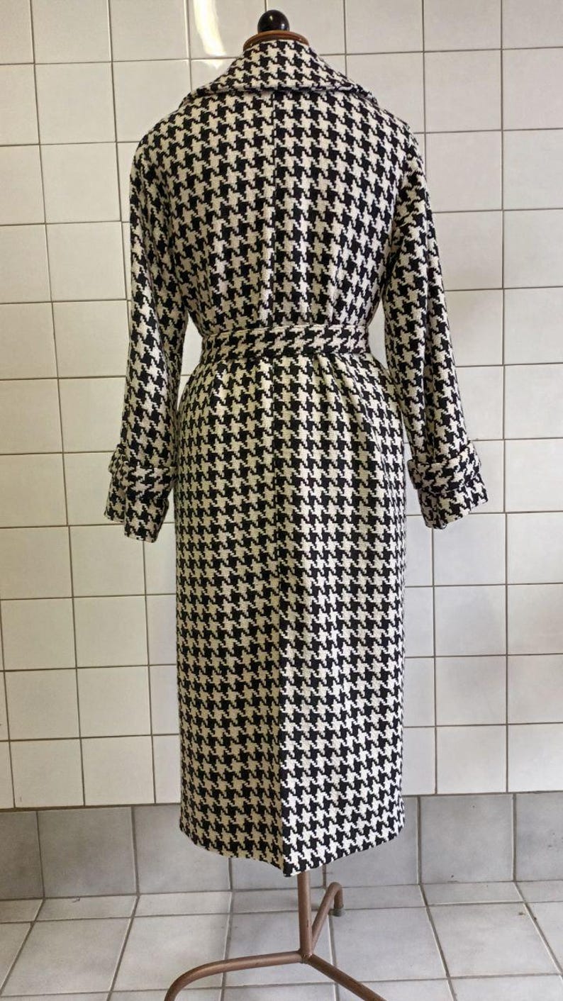 May include: A long, houndstooth-patterned coat in black and cream. The coat features a belted waist, long sleeves with buttoned cuffs, and a classic collar. The coat is displayed on a mannequin.