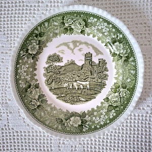 May include: A white ceramic plate with a green floral and landscape design. The centre features a pastoral scene with cows and a castle. The rim has a scalloped edge and is decorated with green floral patterns.