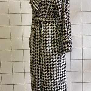 May include: A long, black and white houndstooth pattern coat with a matching belt. The coat has a wide collar, long sleeves, and two front pockets. The coat is displayed on a mannequin against a white tiled wall.
