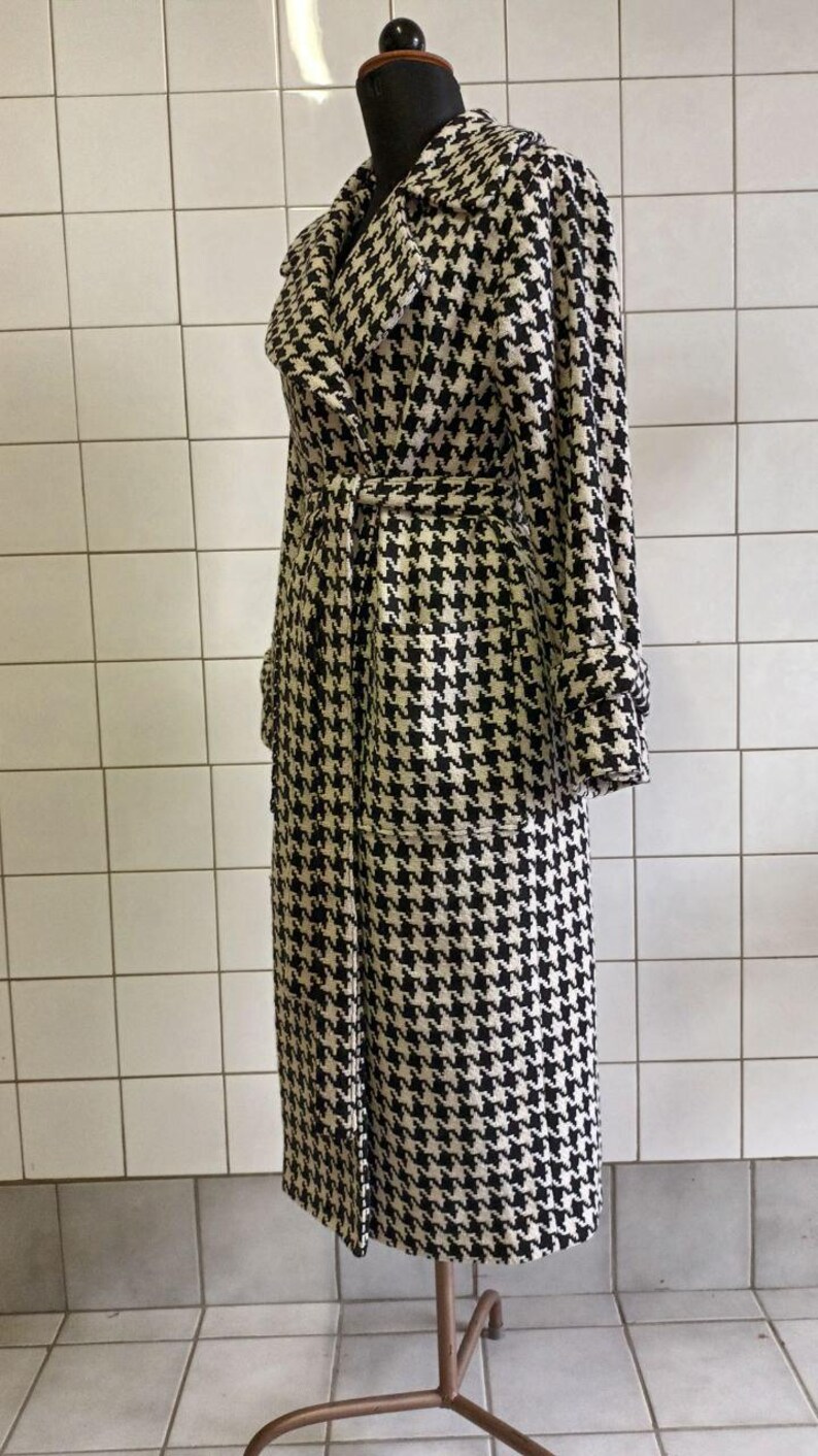 May include: A long, black and white houndstooth pattern coat with a matching belt. The coat has a wide collar, long sleeves, and two front pockets. The coat is displayed on a mannequin against a white tiled wall.