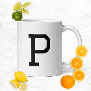 Può includere: Una tazza in ceramica bianca con una lettera "P" nera stampata sul davanti. La tazza è circondata da fette di limoni e lime.