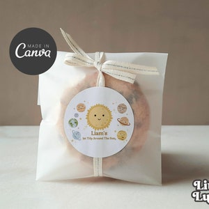 May include: A white paper bag with a white ribbon tied around it. The bag contains a cookie with a round sticker on it. The sticker has a cartoon sun with a smiling face and the text "Liam's 1st Trip Around The Sun".