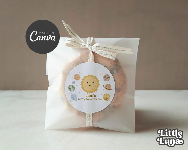 May include: A white paper bag with a white ribbon tied around it. The bag contains a cookie with a round sticker on it. The sticker has a cartoon sun with a smiling face and the text "Liam's 1st Trip Around The Sun".