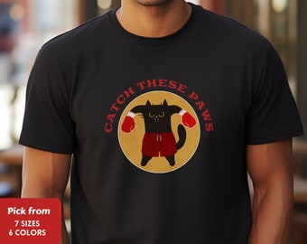 Puede incluir: Camiseta negra con un gato de dibujos animados con guantes de boxeo y pantalones cortos rojos, con el texto "CATCH THESE PAWS" en un diseño circular. El gato está en posición de combate sobre un fondo dorado. Disponible en 7 tallas y 6 colores.