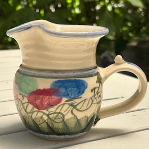 Könnte beinhalten: Ein kleiner, cremefarbener Keramik-Krug mit einem blauen, grünen und roten Blumendesign. Der Krug hat einen Henkel und einen Ausguss.
