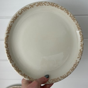 May include: Cream-colored plate with a decorative tan border. The plate is held by a hand, showcasing the smooth surface and the intricate design around the rim. A second plate is partially visible in the lower left corner.
