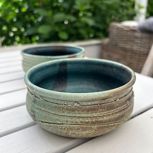 Peut inclure: Deux bols en céramique avec un glaçage vert et des accents bruns. Les bols ont une surface texturée et sont posés sur une table en bois blanc.
