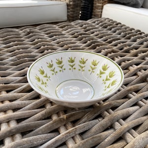 May include: A white ceramic bowl with a green and brown floral pattern. The bowl is sitting on a woven wicker surface.