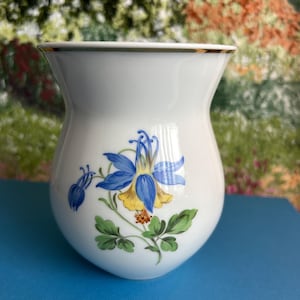 Może przedstawiać: Biały ceramiczny wazon ze złotą obwódką i kwiatowym wzorem. Wzór przedstawia niebiesko-żółty kwiat z zielonymi liśćmi. Wazon stoi na niebieskiej powierzchni.
