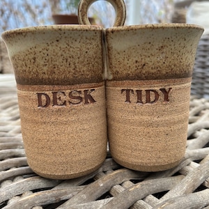 Może przedstawiać: Ręcznie robiony, nakrapiany ceramiczny organizer na biurko z dwoma przegródkami i uchwytem. Na przodzie wytłoczono słowa "DESK TIDY". Ceramika jest w kolorze jasnobrązowym z ciemniejszymi brązowymi akcentami.