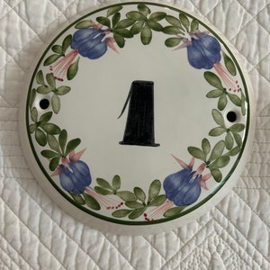 May include: Circular ceramic house number plaque featuring the number 1 in black. The white plaque is decorated with a floral border of blue flowers and green leaves. Includes two holes for mounting.
