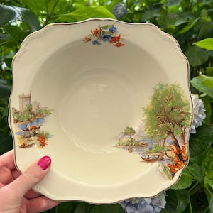 Pode incluir: Uma tigela de cerâmica branca com uma borda ondulada e uma borda dourada. A tigela apresenta uma cena pintada à mão de um lago com um castelo, árvores e barcos. A cena é cercada por uma borda de flores vermelhas, azuis e brancas.