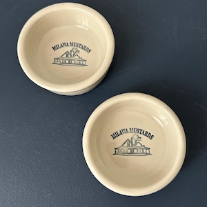 Puede incluir: Dos cuencos de cerámica blanquecina con el texto "Milawa Mustards" y un gráfico azul de un edificio. Los cuencos son redondos con un borde ligeramente elevado. Están sobre una superficie azul oscuro.