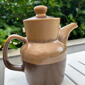 May include: A brown ceramic teapot with a lid and a handle. The teapot has a light brown top and a darker brown bottom.