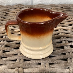 Könnte beinhalten: Ein kleiner brauner und cremefarbener Keramik-Milchkännchen mit Henkel. Der Milchkännchen hat eine braune Oberseite und eine cremefarbene Unterseite.