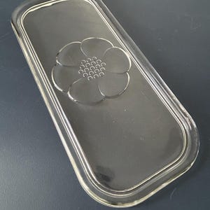 May include: A clear, rectangular glass lid with rounded corners. The lid has a raised floral design in the centre, with a flower shape and small circles. The glass is transparent.
