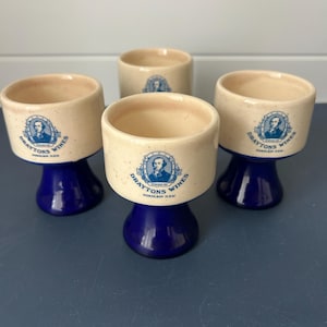 May include: Four vintage wine glasses. Each glass has a cream-colored bowl with a blue base. The glasses feature a blue and white emblem with the text "Draytons Wines" and a portrait.