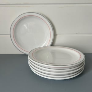 May include: A stack of six white ceramic plates with a grey and red border. One plate is standing upright behind the stack. The plates are on a grey surface with a white background.