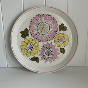 Op de afbeelding: Een witte keramische plaat met een roze en geel bloemmotief. De plaat heeft een roze rand en een patroon van roze, gele en roze bloemen met groene bladeren. De plaat is rond.