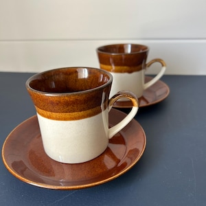 May include: Two ceramic coffee cups and saucers. The cups are white with a brown band and rim, and a brown handle. The saucers are brown. The set is likely vintage or retro, and suitable for serving coffee or tea.