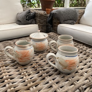 May include: Four white ceramic mugs with a floral design and a white ceramic sugar bowl with a lid on a wicker table.