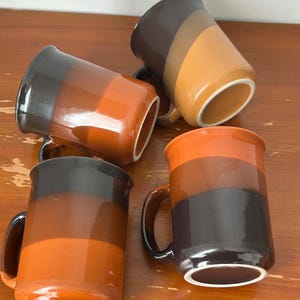 May include: Four ceramic mugs with a colour-blocked design in shades of brown and orange. Each mug has a handle and a white rim. The mugs are arranged on a wooden surface.