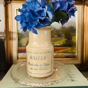 Puede incluir: Un jarrón de cerámica beige con texto azul y un diseño floral, lleno de vibrantes hortensias azules. El jarrón está sobre un plato de vidrio transparente, con una pintura enmarcada en el fondo.