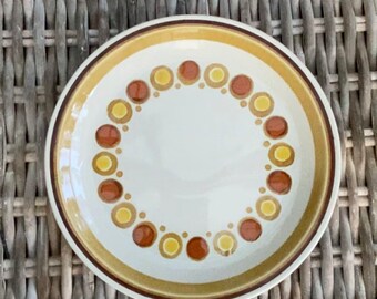 May include: A white ceramic plate with a brown and yellow circle pattern around the edge. The plate is on a woven wicker surface.