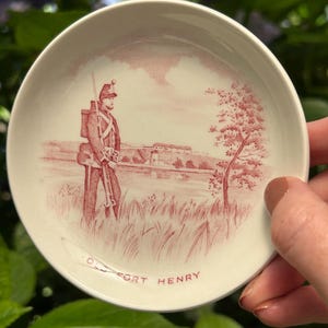 Puede incluir: Un platillo de cerámica blanco con una ilustración rosa y blanca de un soldado de pie frente a un fuerte. El platillo tiene las palabras "Old Fort Henry" impresas en la parte inferior.
