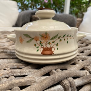 May include: A white ceramic casserole dish with a lid. The dish features a floral design with orange and green flowers and a brown basket.