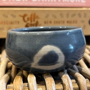 May include: A small, round, blue ceramic bowl with a white, eye-shaped design. The bowl has a glossy finish and sits on a woven wicker surface. The bowl is likely used for serving or display.