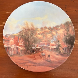 May include: A decorative plate featuring a landscape painting of a town with red brick buildings, trees, and a dirt road. The plate has a light blue sky and is signed "Chris Huber".