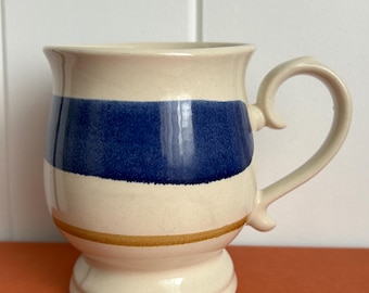 May include: Cream-colored ceramic mug with a blue stripe and a gold accent band. The mug has a curved handle and a pedestal base. The mug is likely handmade and suitable for hot beverages.