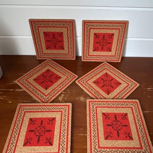 May include: A set of six cork coasters with a red floral design. The coasters have a square or diamond shape, with a brown border featuring a repeating pattern. The central red design is floral.