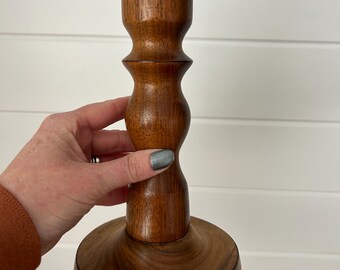 Puede incluir: Un candelero de madera con acabado marrón oscuro. El candelero tiene una base redonda, una sección media bulbosa y una parte superior cilíndrica. La veta de la madera es visible. El soporte está diseñado para una sola vela.