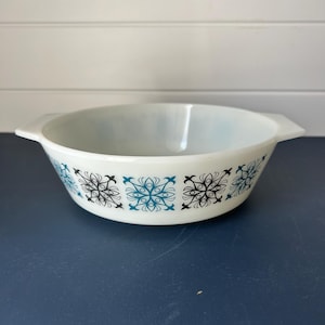 Puede incluir: Fuente ovalada blanca con asas, con un diseño floral en negro y turquesa. La fuente de vidrio está sobre una superficie azul oscuro. Un artículo de cocina vintage, ideal para hornear y servir.