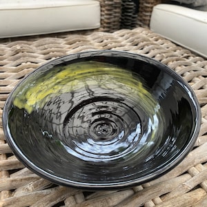 Puede incluir: Un cuenco de cerámica negro con un glaseado amarillo en el interior. El cuenco tiene un acabado liso y brillante y una forma ligeramente redondeada.
