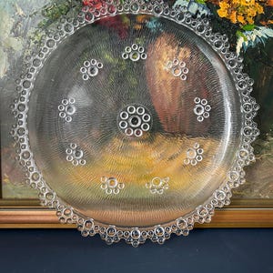 May include: A clear glass plate with a textured surface and a decorative bubble-like edge. The plate has a central design of concentric circles and smaller bubble clusters. The plate is round and appears to be vintage. The diameter is approximately 25 cm.