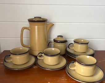 以下が含まれることがあります： 茶色のトリムが付いた、ヴィンテージのベージュセラミックコーヒーセット。セットには、コーヒーポット、砂糖入れ、カップとソーサーがいくつか含まれています。コーヒーセットは木製の表面に展示されています。