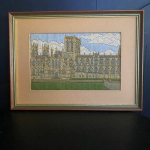 May include: A framed needlepoint embroidery depicting a large, detailed building with a stone facade and a green lawn in front. The building has a pointed roof and many windows. The sky is blue with white clouds.