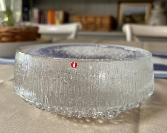 May include: A clear glass bowl with a textured, vertical-line pattern. The bowl has a rounded base with small, bubble-like details. A red and white logo is visible on the side. The bowl is sitting on a beige tablecloth.