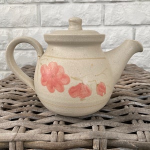 May include: A beige ceramic teapot with a floral design. The teapot has a rounded body and a lid with a knob. The floral design features pink flowers and leaves.