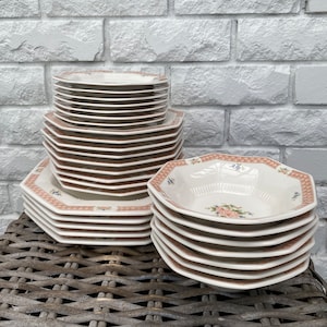 Puede incluir: Una pila de platos de cerámica octogonales blancos con un patrón floral y un borde marrón. Los platos están apilados sobre una superficie de mimbre tejido.