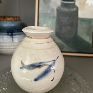 May include: A small, cream-coloured ceramic vase with a rounded form and a narrow neck. The vase features blue and brown leaf-like patterns. Another blue and white vase is visible in the background, along with a painting of flowers.