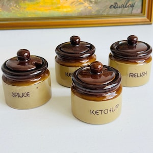 May include: A set of four small ceramic condiment jars with dark brown lids and tan bases. Each jar features a dark brown knob on the lid and black text printed on the side. The labels read Sauce, Ketchup, and Relish, with one jar partially obscured.